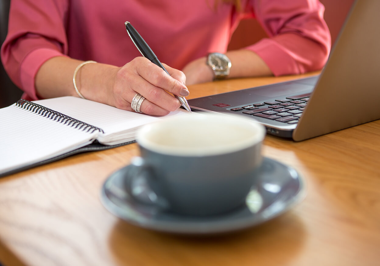 Someone writing on a notepad with a cup and laptop on the desk