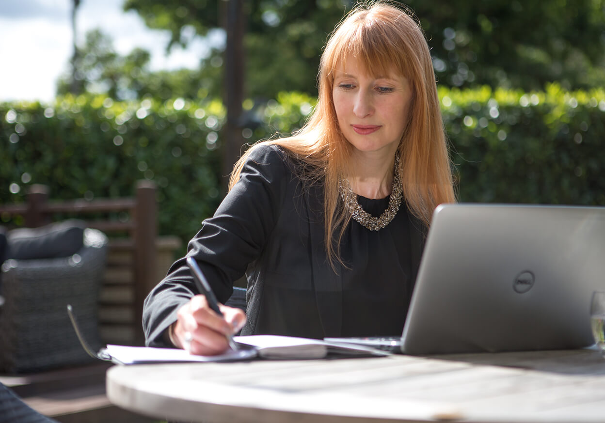 Woman outside writing on a notepad with a laptop on the table