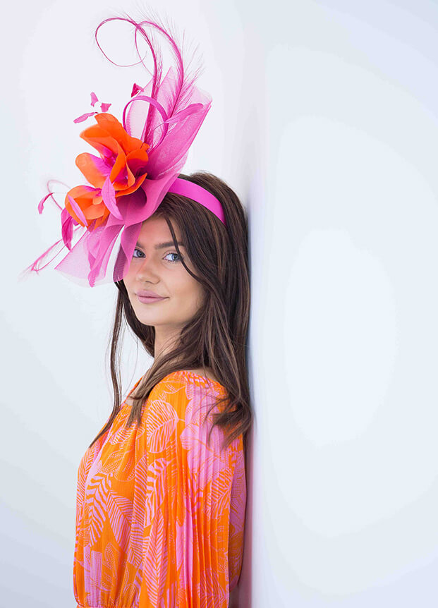 Woman standing next to a wall in a orange and pink dress wearing a flamboyant hat
